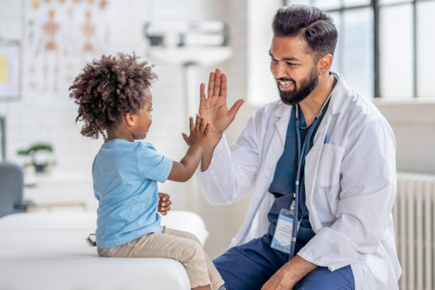 A sweet little boy of African decent sits up on an exam table during a doctors appointment.  He is dressed casually and is giving the doctor a high-five at the end of the visit.  The doctor is wearing blue scrubs, a white lab coat and has a stethoscope around his neck as he smiles at the young boy.